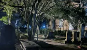 A nighttime view of an old cemetery with weathered gravestones, lit by ambient lights that cast shadows among the trees.