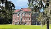 A traditional red brick mansion with a red roof stands amidst a lush green lawn, partially shrouded by the Spanish moss-draped branches of tall trees under a clear blue sky.