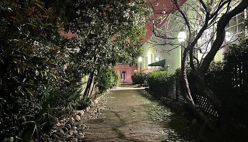 The image depicts a night scene of a cozy cobblestone pathway flanked by lush greenery and warm street lamps leading towards a building entrance