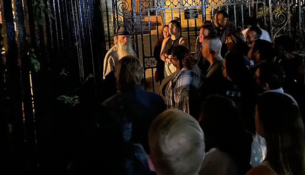 A group of people is gathered at night around an entrance where a person with a white beard appears to be speaking or performing