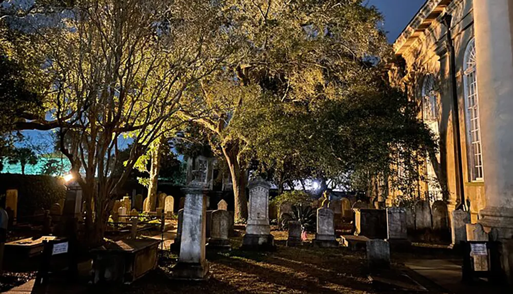 The image depicts an old cemetery at night bathed in artificial light that casts an eerie glow on the historic gravestones and surrounding trees