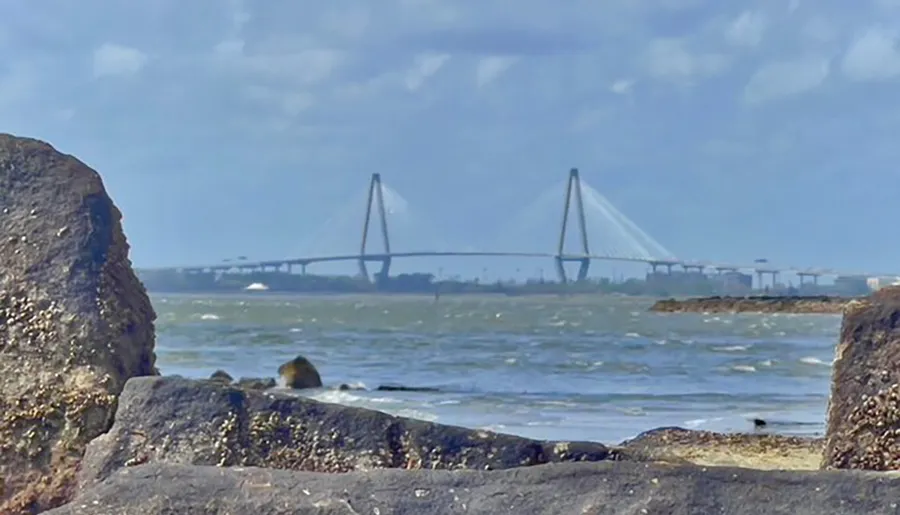 The image shows a coastal scene with rough sea waters in the foreground and a modern cable-stayed bridge in the background, framed by rocky outcroppings.