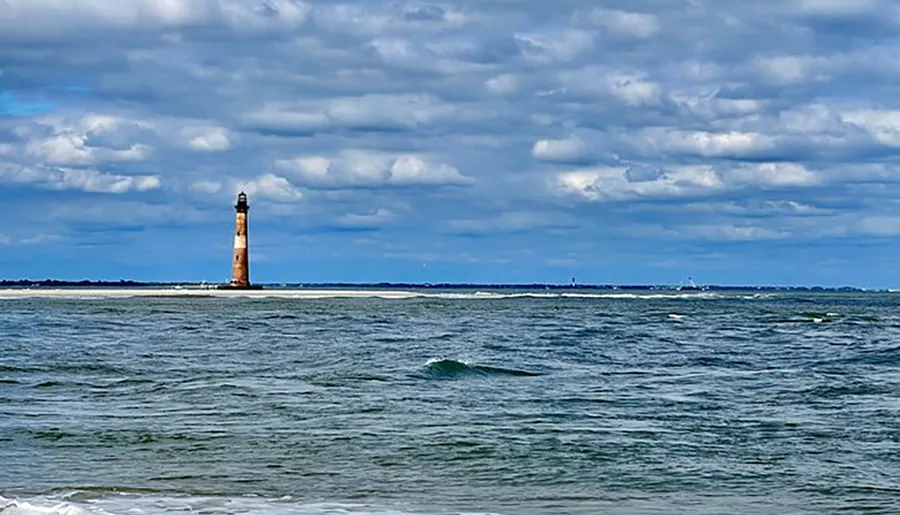 A solitary, weathered lighthouse stands tall on a narrow strip of land between a cloudy sky and the choppy waters of the sea.