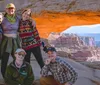 A group of five people pose for a photo under a natural arch with a panoramic view of canyons in the background