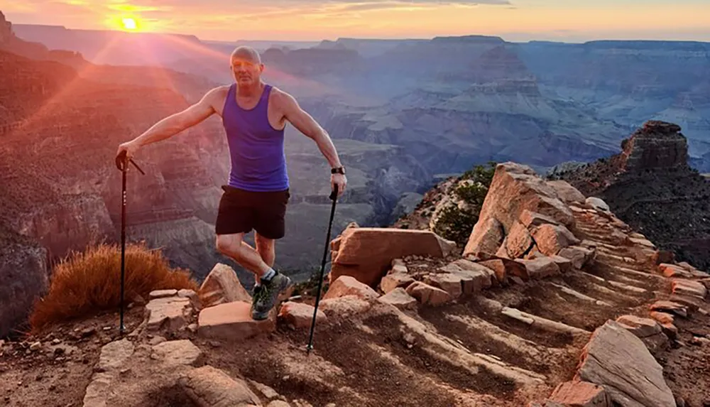 A person stands triumphantly atop a rocky trail with a trekking pole overlooking the Grand Canyon during a breathtaking sunset