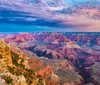 A person in a red and black plaid shirt stands in front of the vast and scenic Grand Canyon under a clear blue sky