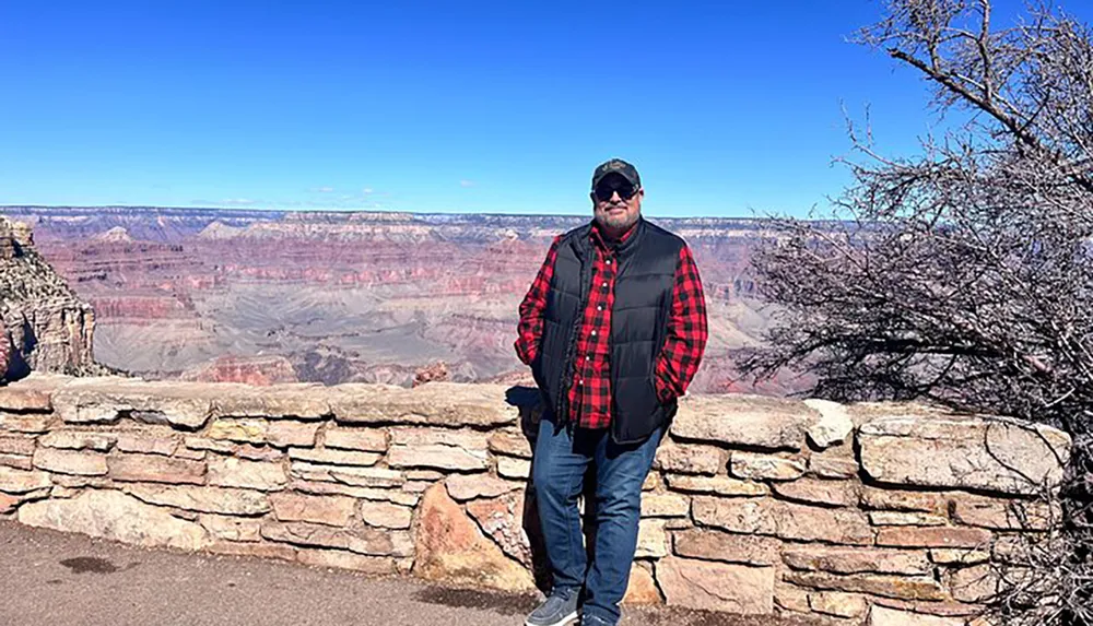 A person in a red and black plaid shirt stands in front of the vast and scenic Grand Canyon under a clear blue sky