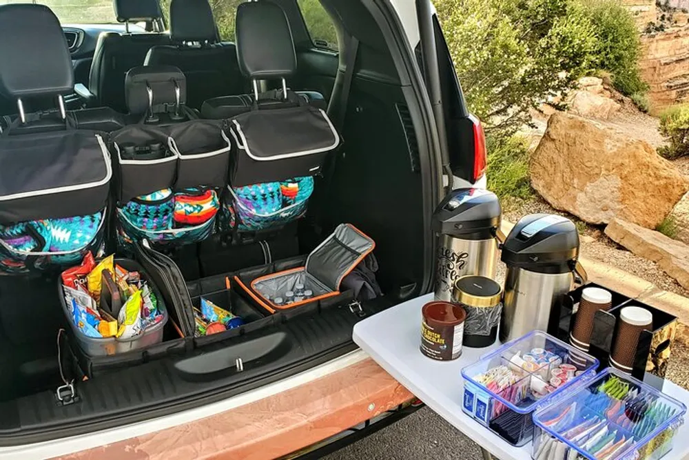 The image shows the open trunk of a vehicle well-organized with various storage compartments containing snacks and supplies and a set-up of thermos flasks and disposable cups suggesting preparation for a road trip or outdoor event