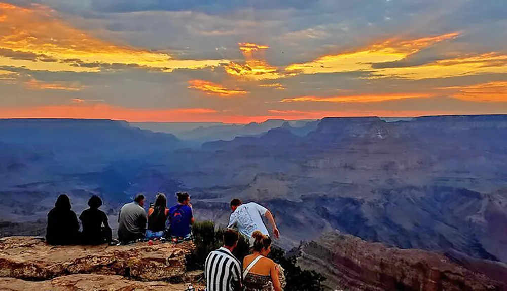 A group of people is sitting and standing on the edge of a canyon enjoying the view of a vibrant sunset