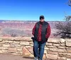 A person is standing in front of a low stone wall with the vast expanse of the Grand Canyon in the background under a clear blue sky