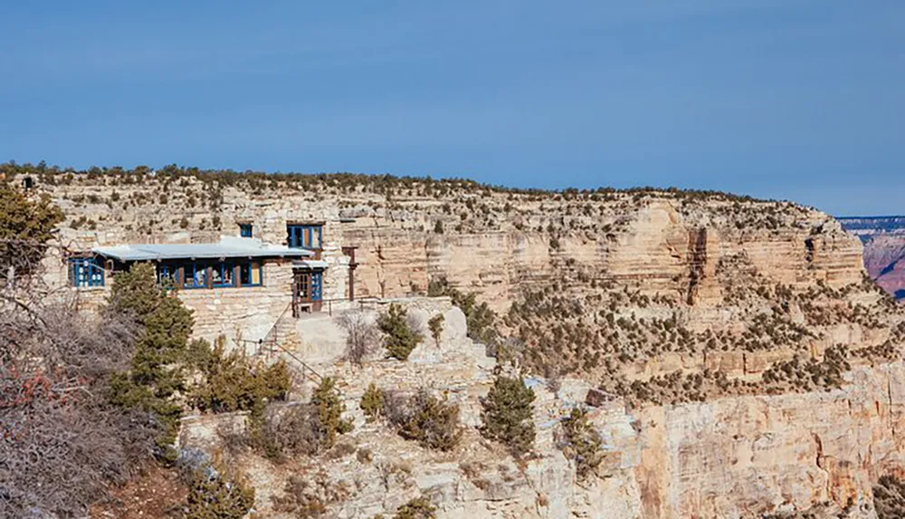 The image shows a stone building with blue window frames perched on the edge of a cliff overlooking a vast canyon landscape under a clear sky