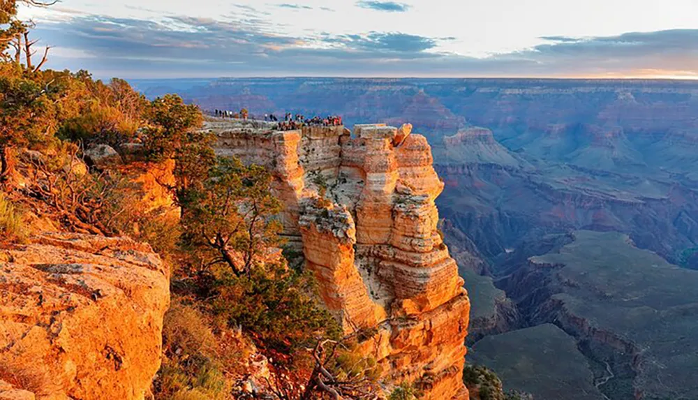 The image captures a group of people standing at the edge of a scenic viewpoint overlooking the vast and colorful expanse of the Grand Canyon at sunset