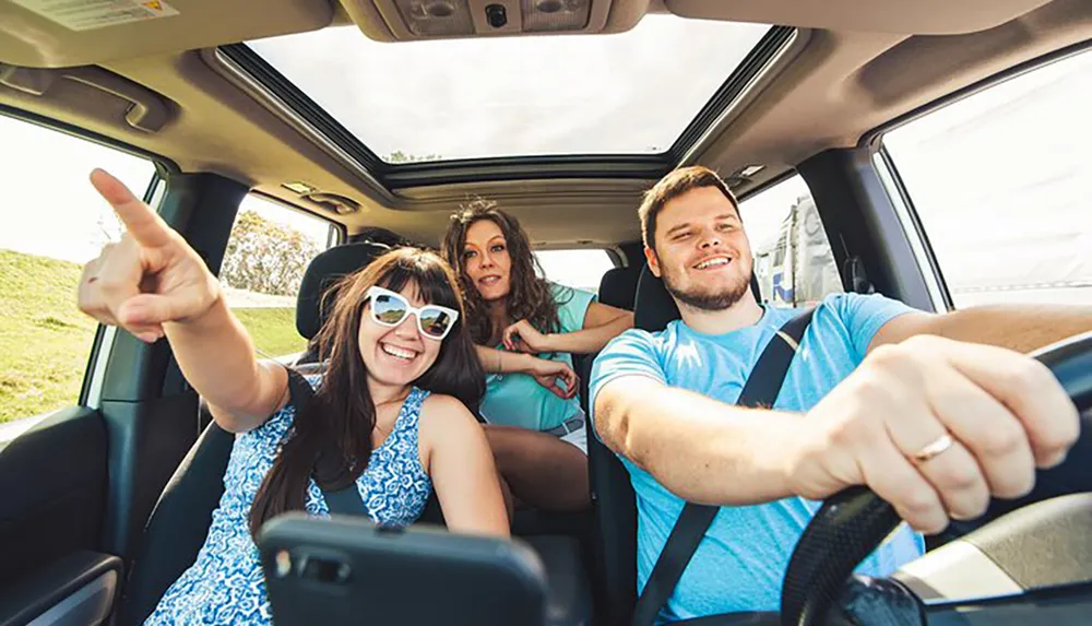 Three cheerful people are taking a selfie in a car with one of them pointing out of the window and the driver focusing on the road