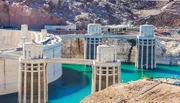 The image shows the Hoover Dam with its distinctive concrete arch-gravity structure, flanked by large water intake towers, situated in a rugged desert canyon with a reservoir behind it.