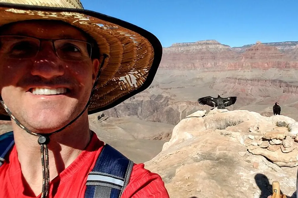 A person in a wide-brimmed hat takes a selfie with the Grand Canyon in the background while another person and a large bird possibly a condor are visible in the distance