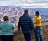 A group of tourists led by a guide in a yellow shirt is observing the vast expanse of the Grand Canyon from a viewpoint behind a safety railing