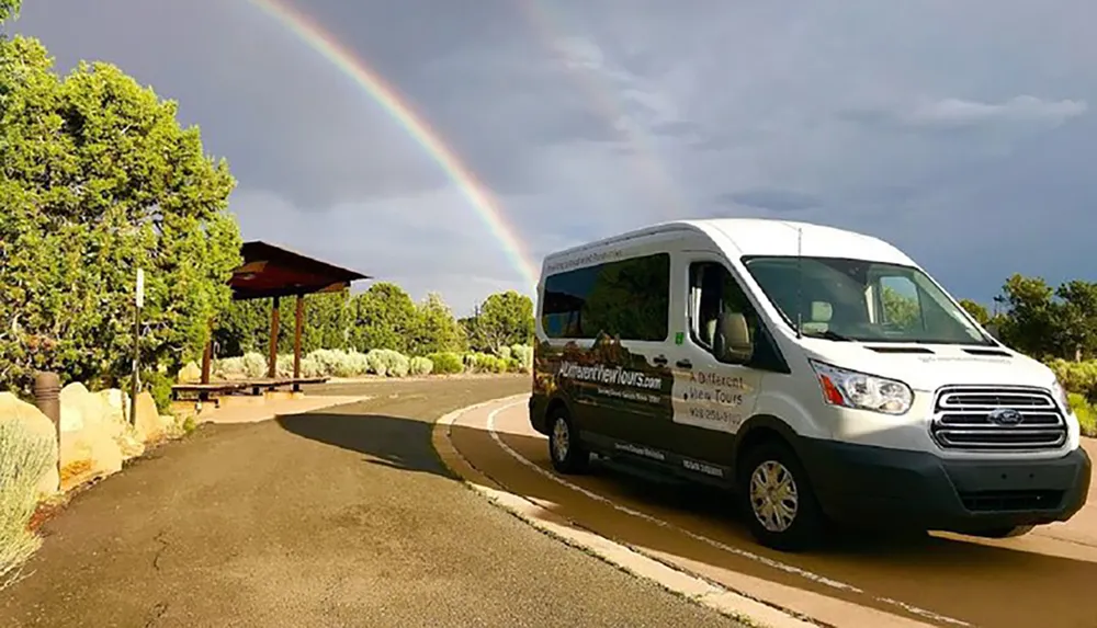 A shuttle van is parked on a sunny road with a vivid rainbow in the background