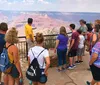 A group of tourists led by a guide in a yellow shirt is observing the vast expanse of the Grand Canyon from a viewpoint behind a safety railing