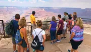 A group of tourists, led by a guide in a yellow shirt, is observing the vast expanse of the Grand Canyon from a viewpoint behind a safety railing.