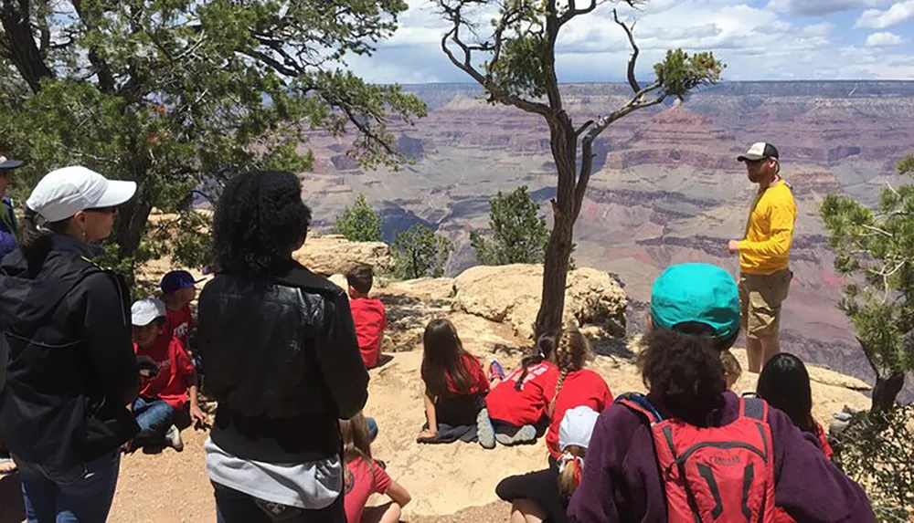 A group of people including children are attentively listening to a guide at the edge of the Grand Canyon