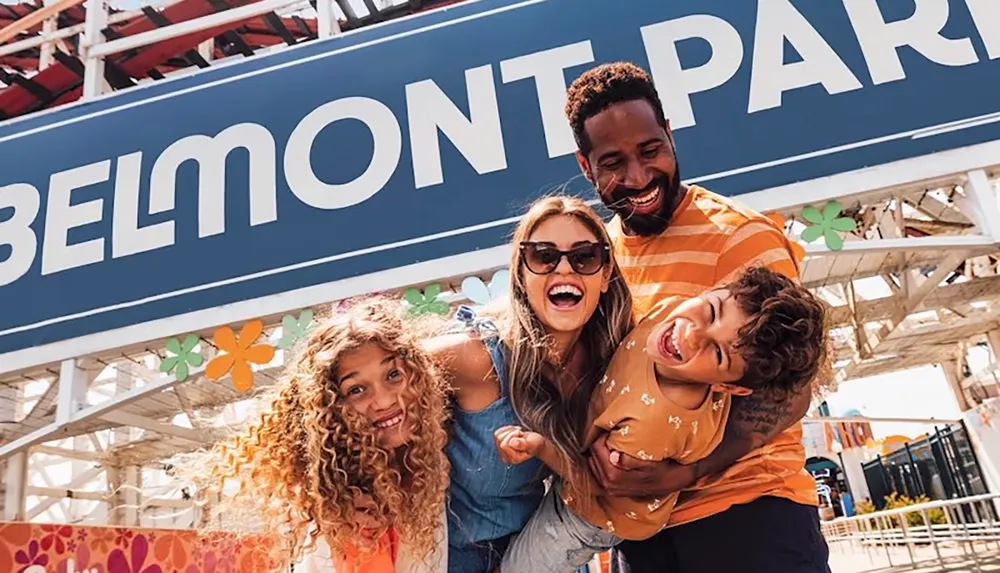 A joyful family is laughing and posing in front of the BELMONT PARK sign at an amusement park