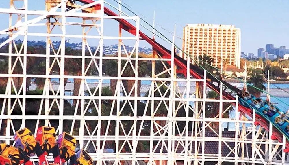 The image shows a wooden roller coaster with a bright cityscape and water in the background