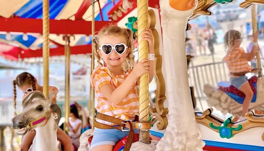 A young girl wearing heart-shaped sunglasses and a striped shirt is joyfully riding a carousel horse