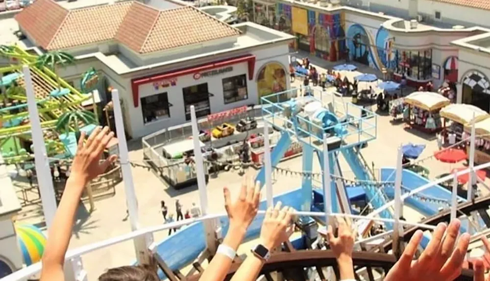 The image shows a persons view from a roller coaster with raised hands overlooking a bustling amusement park area with rides and crowds