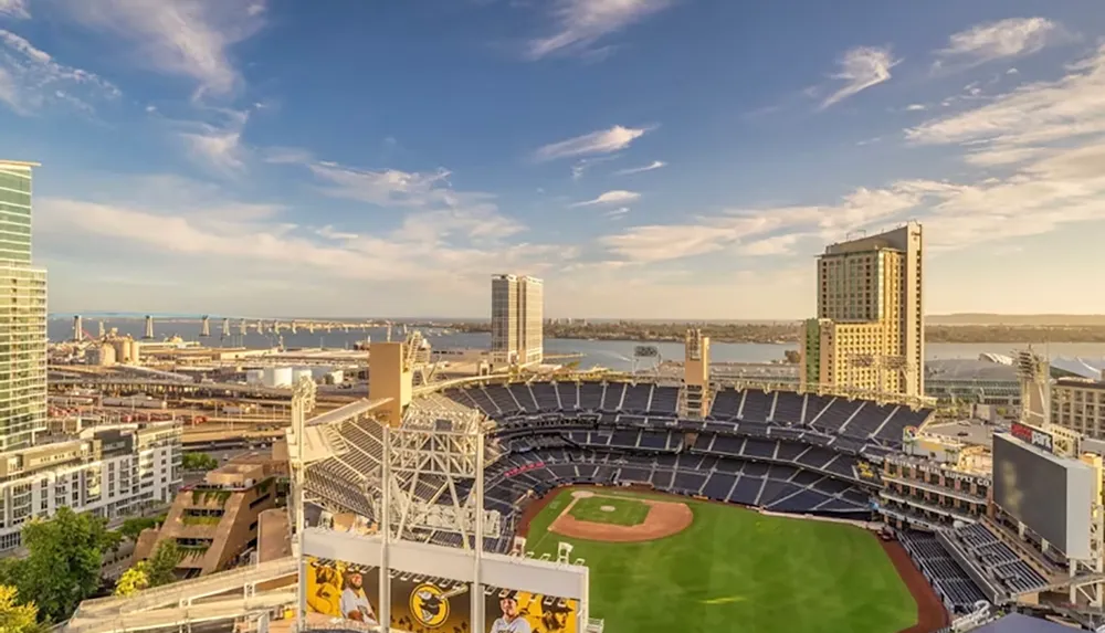 The image shows a panoramic view of a baseball stadium surrounded by city buildings and water under a partly cloudy sky