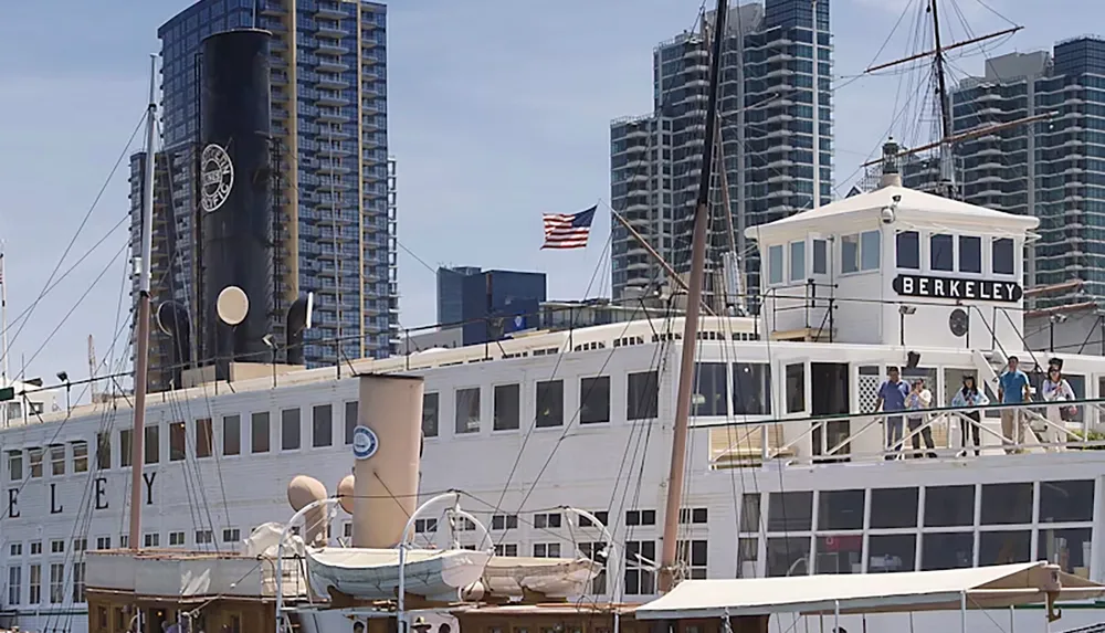 The image depicts the historic steam ferry Berkeley docked with several people aboard set against a backdrop of modern high-rise buildings and an American flag