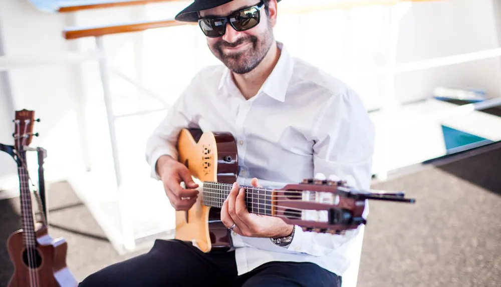 A man wearing sunglasses and a hat is smiling while playing a guitar