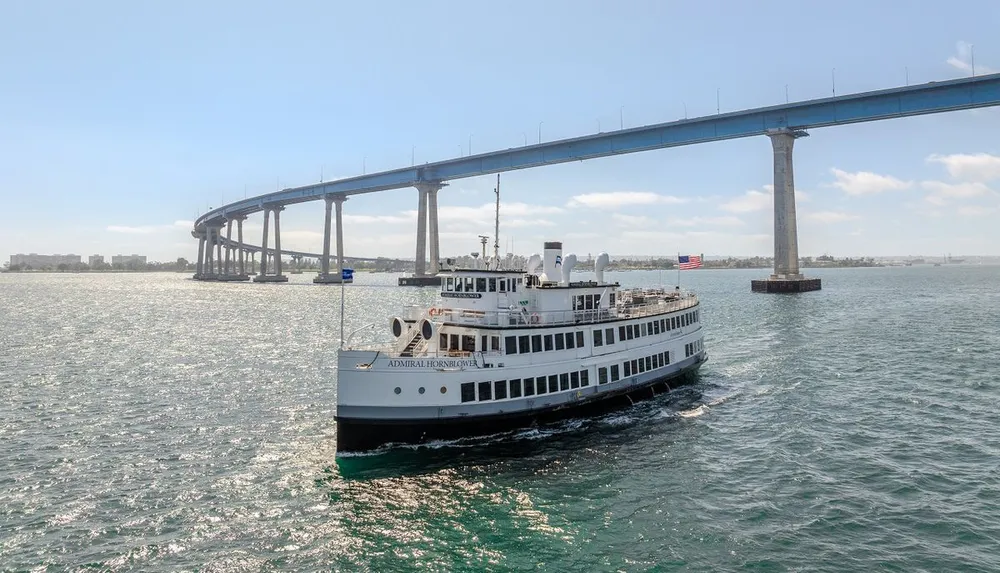 A white passenger ship sails under a large arched bridge on a sunny day