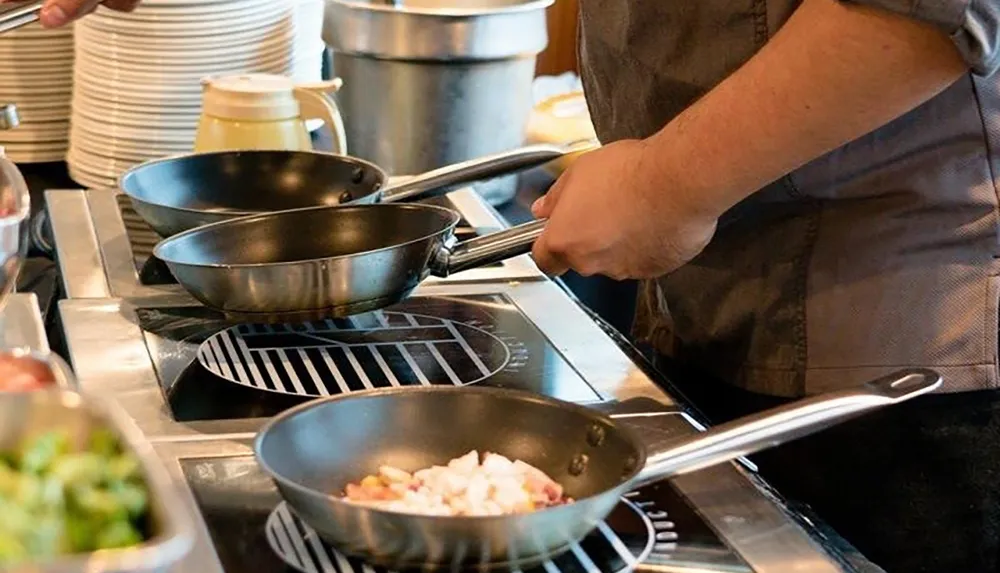 A person is cooking with multiple frying pans on a stove in a kitchen setting