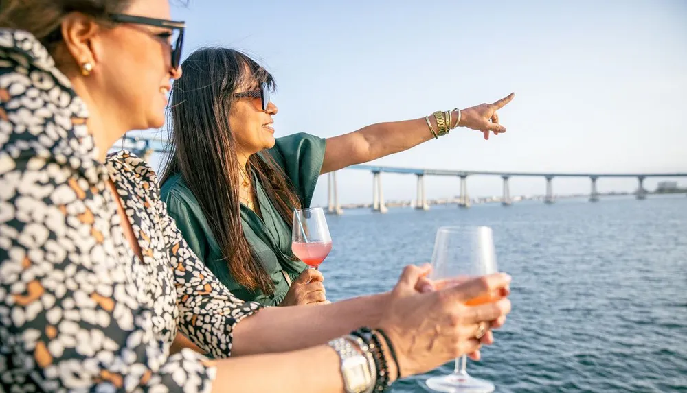 Two women are enjoying drinks on a boat while one of them points towards something in the distance over the water