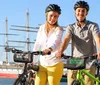 Two people wearing helmets are standing with their bikes near a waterfront with a sailing ship in the background