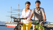 Two people wearing helmets are standing with their bikes near a waterfront, with a sailing ship in the background.