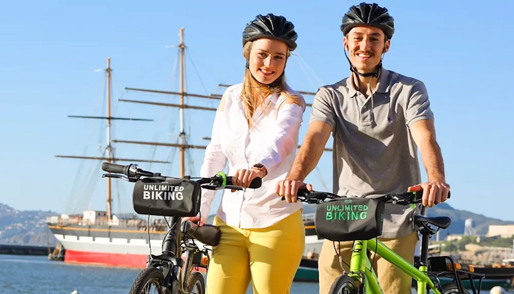 Two people wearing helmets are standing with their bikes near a waterfront with a sailing ship in the background