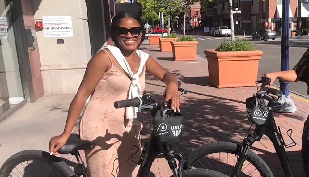 A woman in sunglasses is smiling and posing with a bicycle on a sunny day in an urban setting