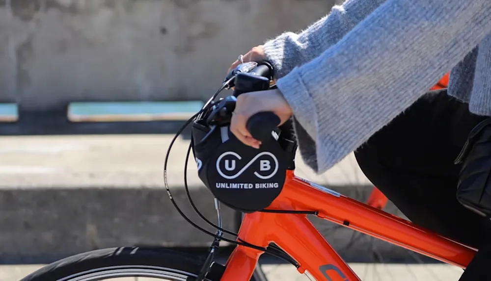 A person wearing a gray sweater is riding an orange bicycle with a Unlimited Biking logo on the handlebars