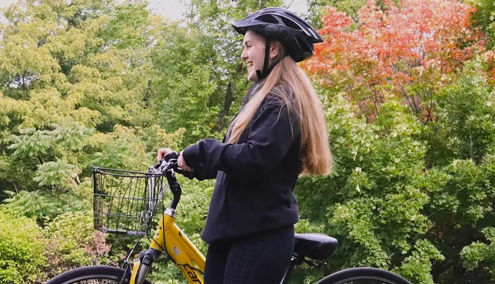 A person wearing a helmet stands next to a yellow bicycle in a park with green and red foliage