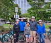 A group of people wearing helmets is gathered with their bicycles in a park surrounded by trees and a building in the background