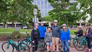 A group of people wearing helmets is gathered with their bicycles in a park, surrounded by trees and a building in the background.