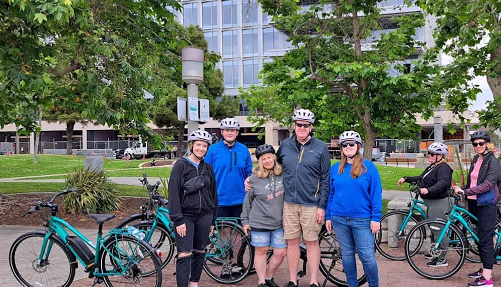 A group of people wearing helmets is gathered with their bicycles in a park surrounded by trees and a building in the background