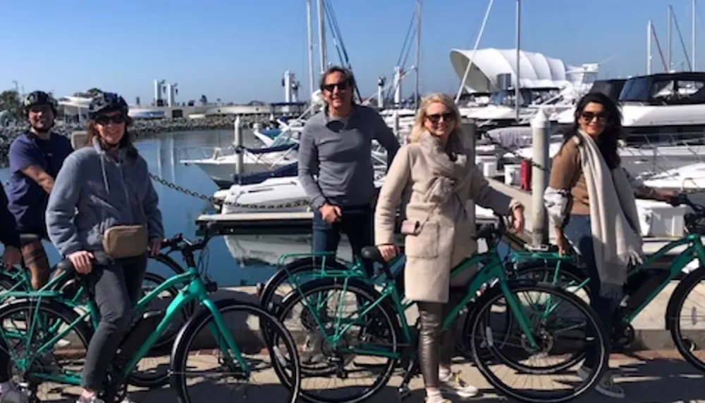 A group of people with bicycles is posing for a photo in front of a marina filled with boats