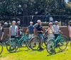 A group of people wearing helmets is gathered with their bicycles in a park surrounded by trees and a building in the background