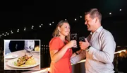 A couple is toasting with wine glasses at a nighttime outdoor setting, with a plated dish of food inset in the image.