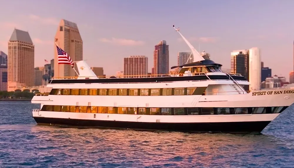 The image shows a white and black cruise ship named Spirit of San Diego sailing on the water with a city skyline in the background and an American flag flying on the ship