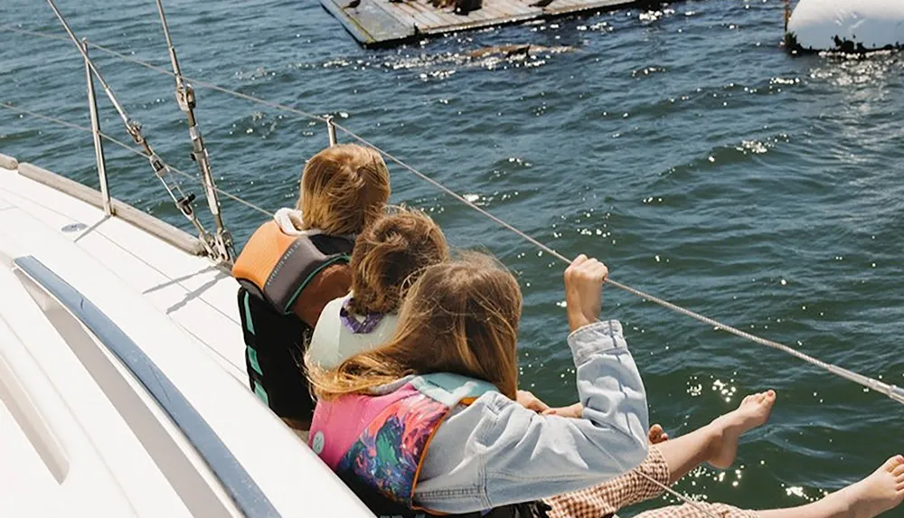 Three people wearing life jackets sit on a sailboat with their feet dangling over the side enjoying the water view