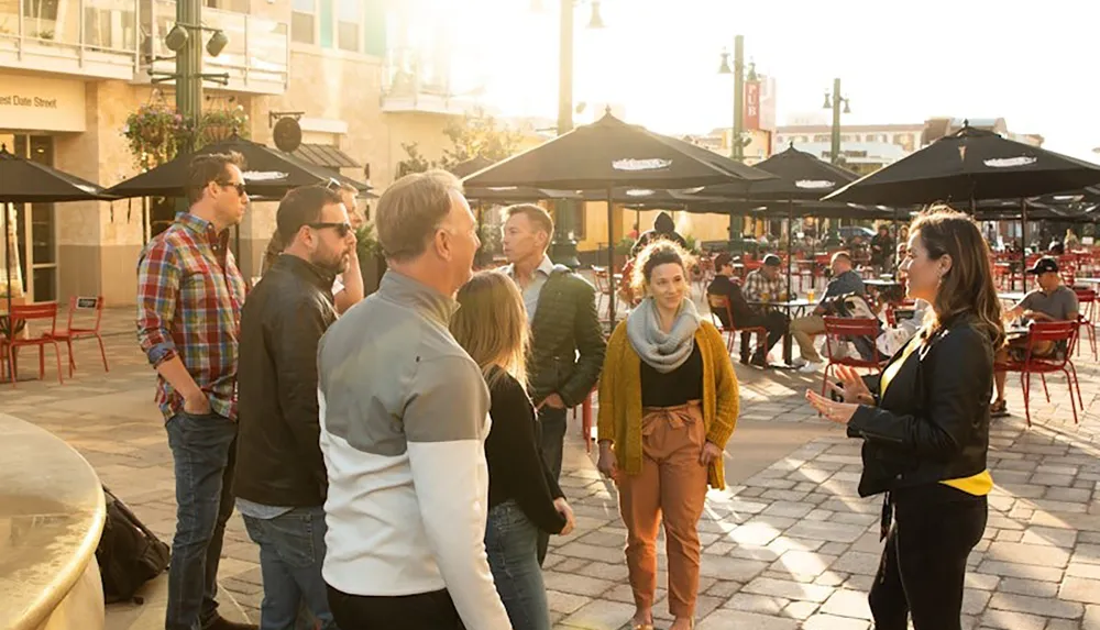 A group of people are gathered outdoors engaged in conversation near a plaza with tables and umbrellas