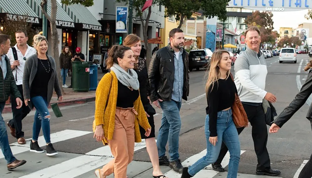 A group of people are happily walking across a street in an urban area with shops and a Little Italy sign in the background